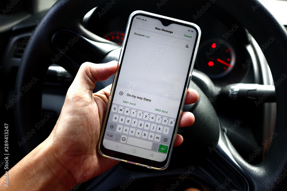 Hand holding a smartphone with a message in front of a car steering wheel Hand holding a smartphone with a message in front of a car steering wheel