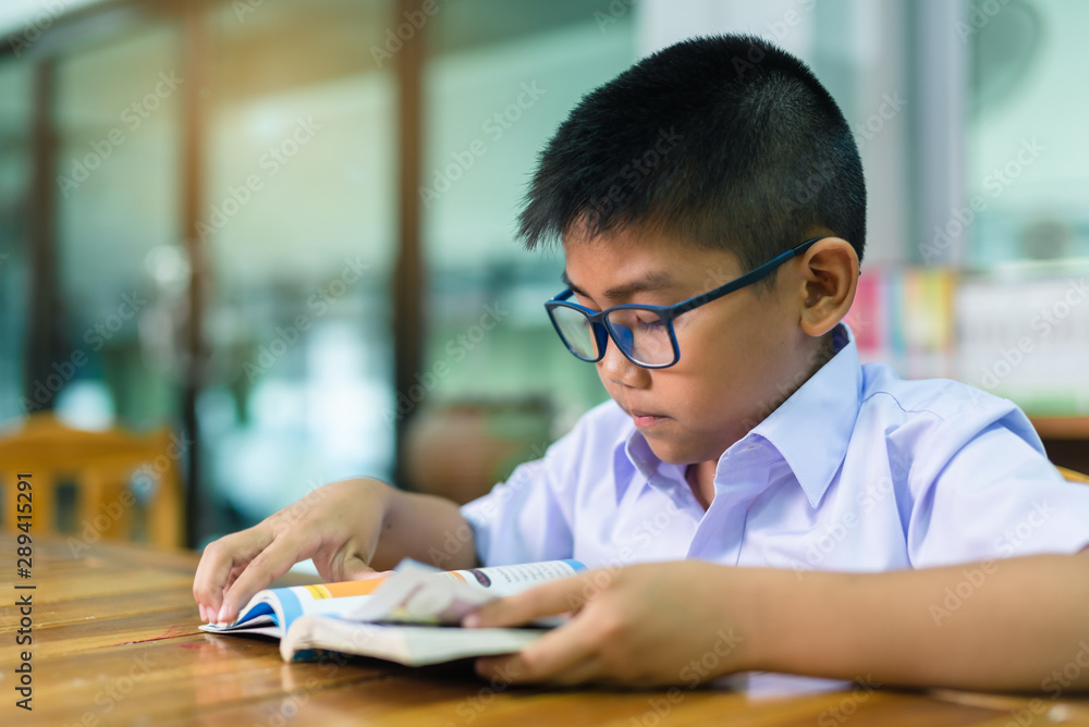 A cute Asian elementary school boy wearing blue glasses in a white school uniform is sitting, enjoying , reading comics in the library. A cute Asian elementary school boy wearing blue glasses in a white school uniform is sitting, enjoying , reading comics in the library.