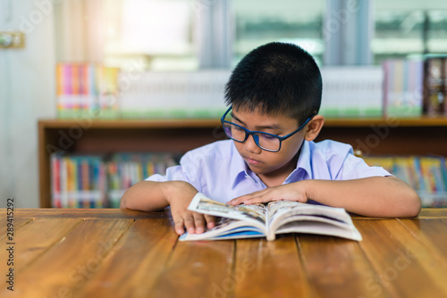 A cute Asian elementary school boy wearing blue glasses in a white school uniform is sitting, enjoying , reading comics in the library.
