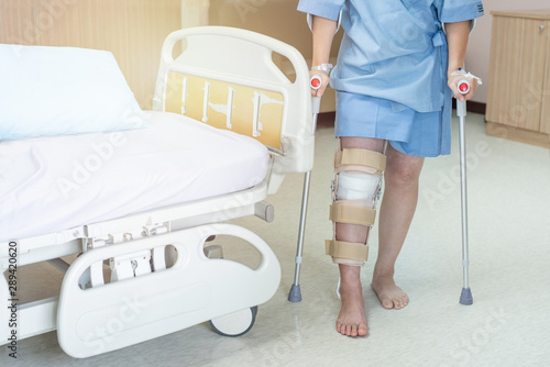 Asian woman patient with knee brace and walking stick in hospital ward after ligament surgery.