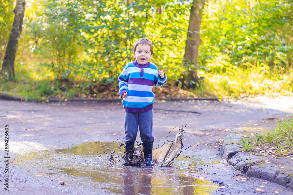 A little boy is jumping in a puddle. A boy in rubber boots. Happy childhood. Puddles after the rain. Warm summer evening. A little boy is jumping in a puddle. A boy in rubber boots. Happy childhood. Puddles after the rain. Warm summer evening.