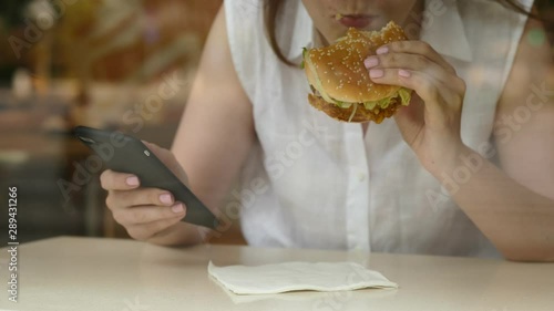 Woman eat chicken burger and watch news feed on smartphone, sit at fast food restaurant, seen through window glass. Lady rise hand and make bite of hamburger