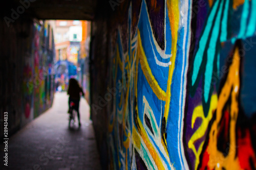Close up of a blue and yellow graffity in Werregarenstraat (Graffiti Street) in Ghent, Belgium, Europe, with defocused background with a person on a bike. Colorful famous street in the old town © Jesus Barroso