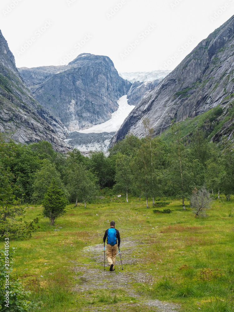 An indefinite man with t goes to the side of a glacier through a valley in Norway. One of the branches of the Nigardsbreen glacier, the view from the valley and the tourist, comes with hiking sticks