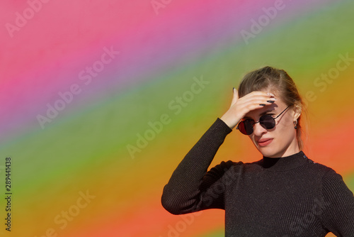 Young attractive woman in sunglasses covers her eyes from the bright sun, bright rainbow background, copy space, close-up. The concept of choice.