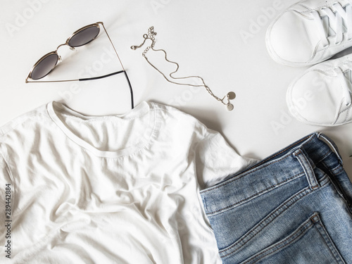 White t-shirt blue jeans, sunglasses, necklace and white sneakers on white background. Flat lay woman's casual outfits. top view