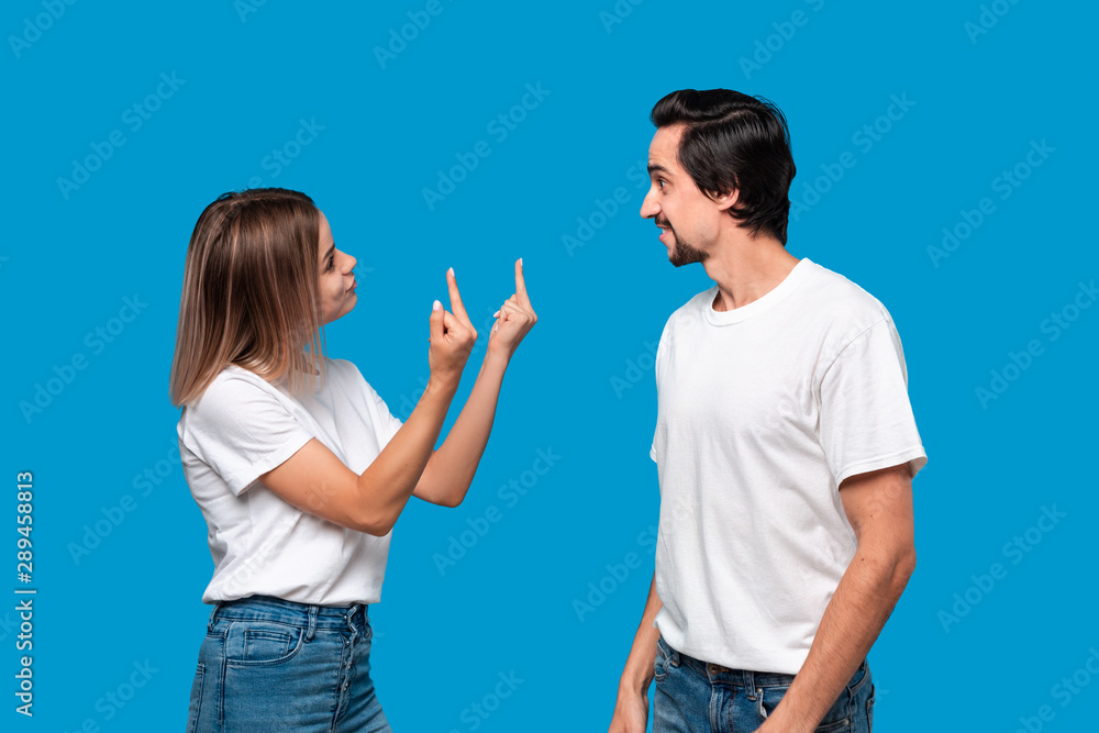 Couple of a young blond woman and brunet bearded man with mustaches in white t-shirts and blue jeans quarreling standing isolated over blue background. Couple of a young blond woman and brunet bearded man with mustaches in white t-shirts and blue jeans quarreling standing isolated over blue background.