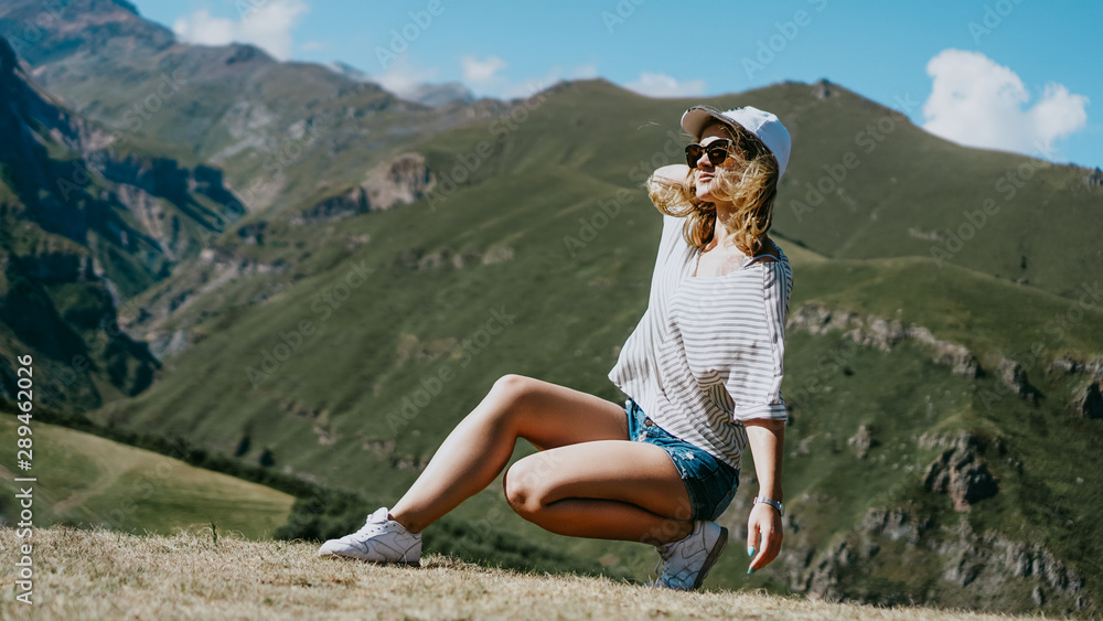 The girl in a cap and shorts in the mountains sits on the grass. Hike in the mountains, resting on a meadow, outdoor