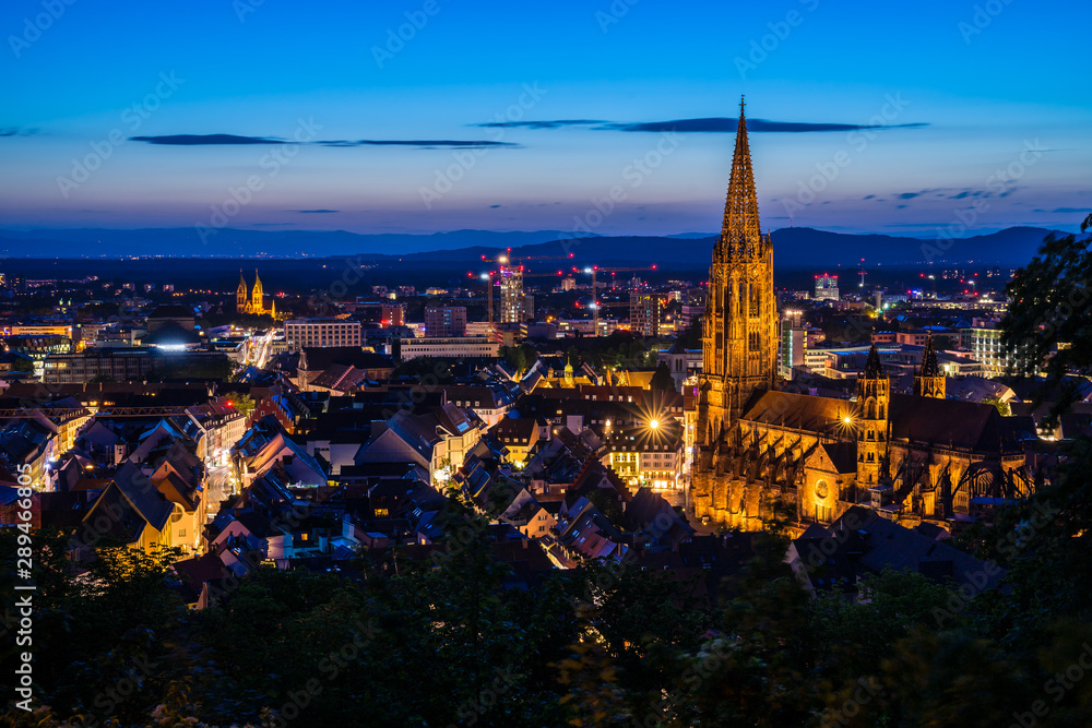 Germany, Freiburg im breisgau city in black forest nature with illuminated ancient historical muenster church surrounded by houses, aerial perspective by night