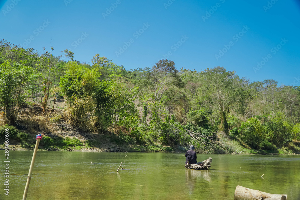 Old man is fishing in the middle of the river while sitting on a tree trunk
