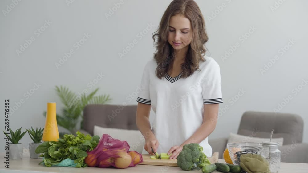 Young slim woman slicing vegetables with the sharp knife at the table in the kitchen. Concept of healthy food. Many fruit and vegetables lying in the foreground Young slim woman slicing vegetables with the sharp knife at the table in the kitchen. Concept of healthy food. Many fruit and vegetables lying in the foreground