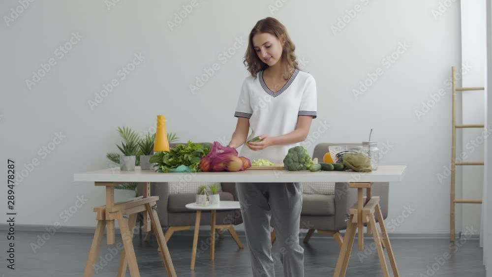 Pretty woman cuts a cucumber on a kitchen cutting board with knife while standing at a table in the kitchen at home. Skillful cook prepares breakfast of vegetables. Vegetable salad. Healthy eating. Pretty woman cuts a cucumber on a kitchen cutting board with knife while standing at a table in the kitchen at home. Skillful cook prepares breakfast of vegetables. Vegetable salad. Healthy eating.