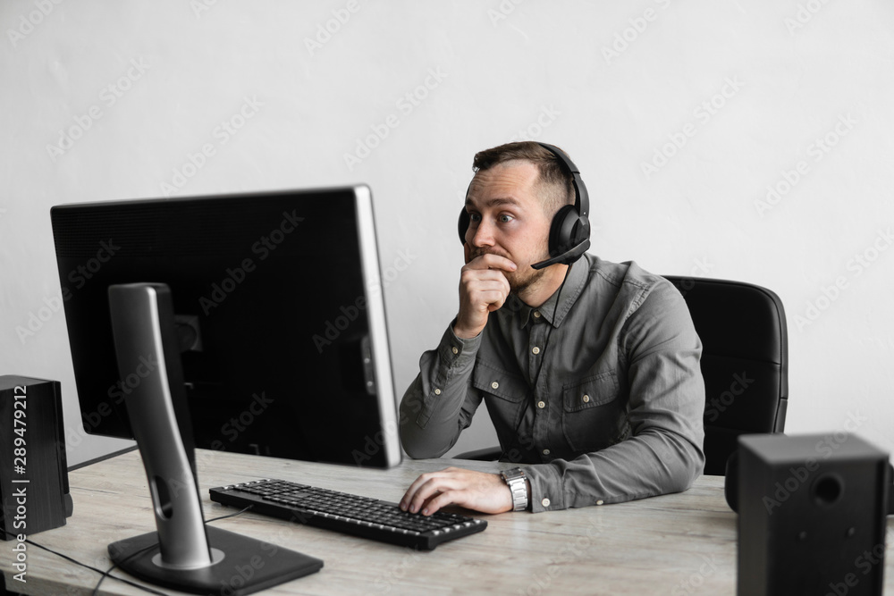 Confused or angry businessman or student in a shirt sitting against monitor of computer in headphones and speaking with somebody via internet. Working on a pc at a table in the office. Confused or angry businessman or student in a shirt sitting against monitor of computer in headphones and speaking with somebody via internet. Working on a pc at a table in the office.