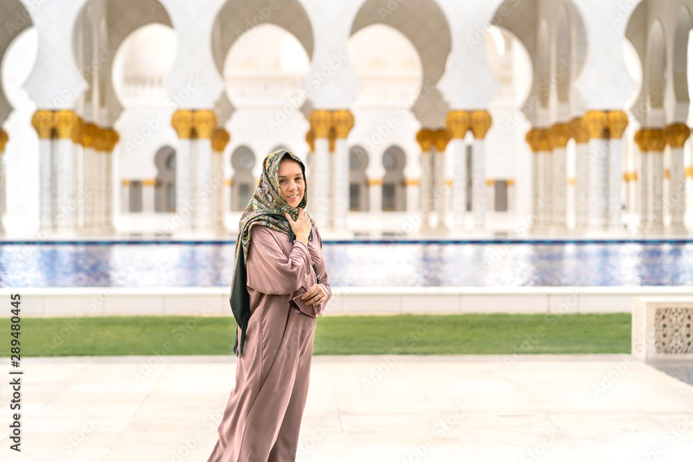 Happy female tourist wearing scarf and dress in The Sheikh Zayed Grand Mosque in Abu Dhabi. Smiling woman walking in beautiful building with water and traditional Muslim decor. Vacation and travel.