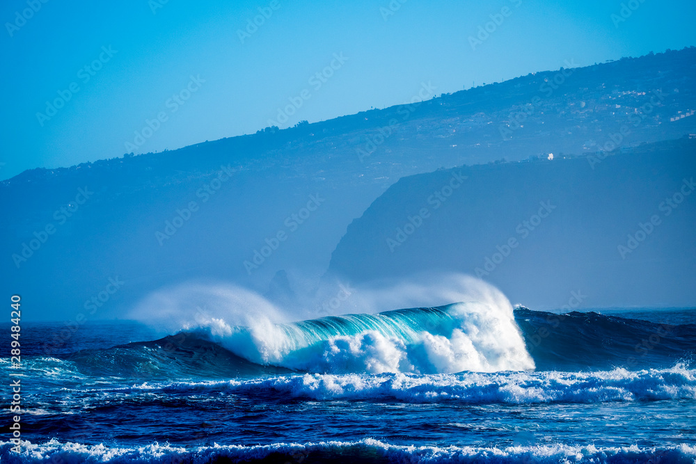 beautiful and bug wave is breaking on the sea or ocean - blue water and pacific or athlantic - houses and mountains at the background - graet place to surf