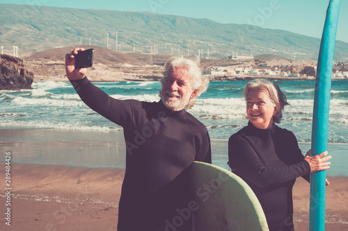 happy couple of seniors at the beach trying to go surf and having fun together - mature woman and man married taking a selfie with the wetsuits and surftables with sea or ocean at the background photo