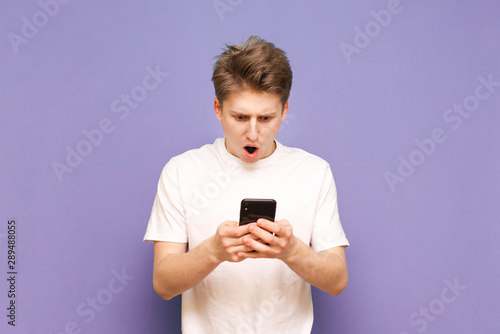 Amazed young man in a white T-shirt uses a smartphone and reacts