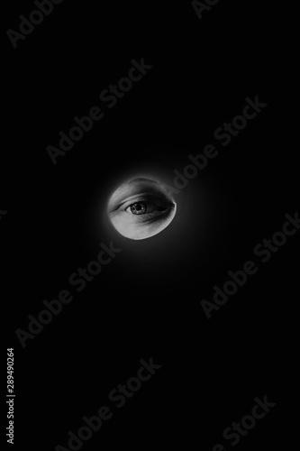 Halloween concept on black background. Eye in the dark. Person looks through the peephole. Creative art idea for scary story. Eye like moon