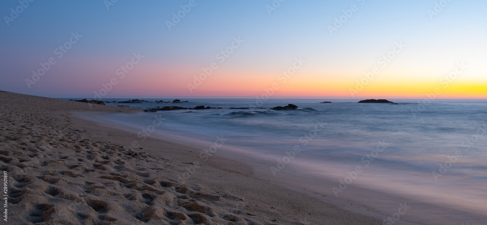 Long exposure shot over beach at dusk with milky ocean and smooth blue orange gradient sky. Dark rocks in water.
