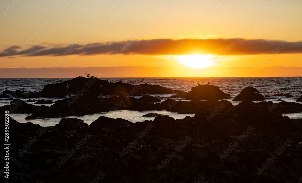 Vivid ocean sunset with dark rocks in silhouette on beach, and sun on horizon