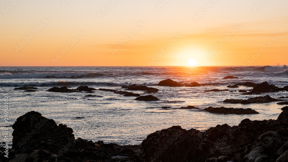 Sunset ocean scene with dark rocks in silhouette and yellow orange gradient sky.