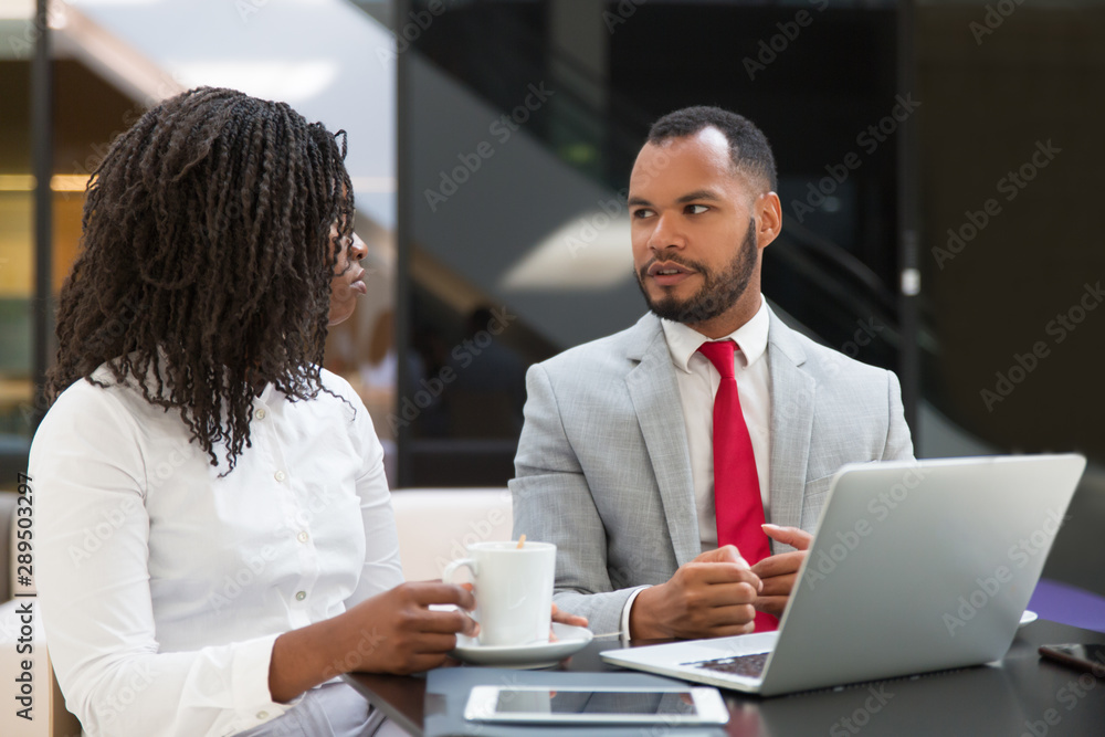 Diverse couple of colleagues watching and discussing content on laptop. Business man and woman drinking coffee, using computer and talking. Digital communication concept Diverse couple of colleagues watching and discussing content on laptop. Business man and woman drinking coffee, using computer and talking. Digital communication concept