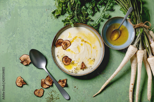 Parsnip cream soup in ceramic bowl with sun dried pears, butter sauce, bundle of fresh parsnip and herbs over green texture background. Flat lay, space