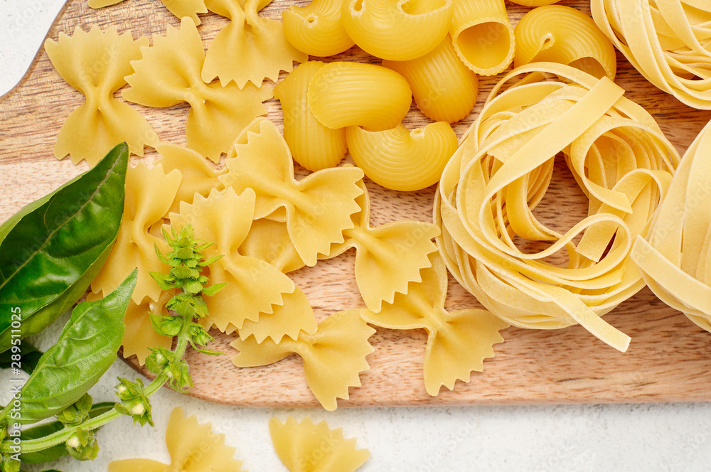 Different types of Italian pasta and fresh basil on wooden cutting board