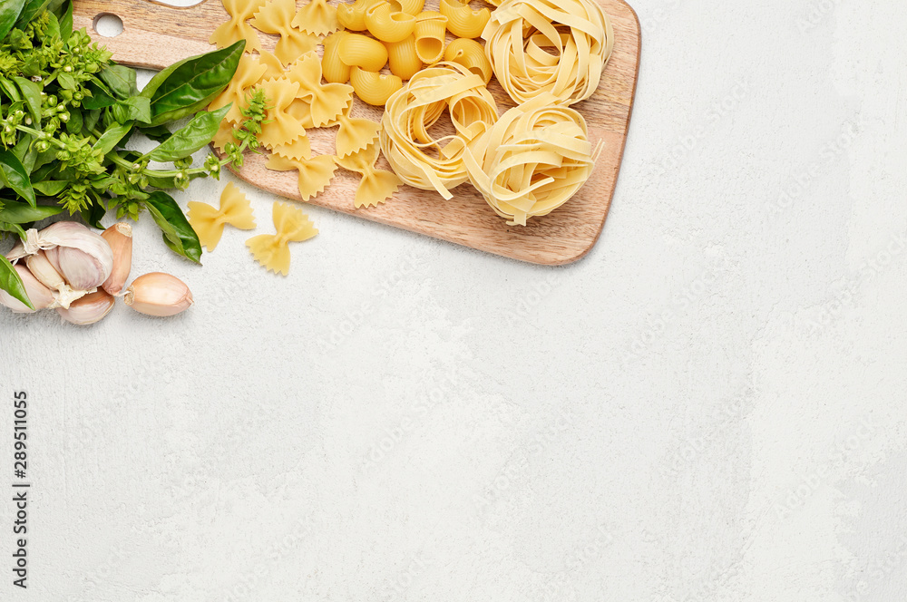 Different types of Italian pasta, fresh basil and garlic. Farfalle, Pipe rigate and Fettuccine on light grey background