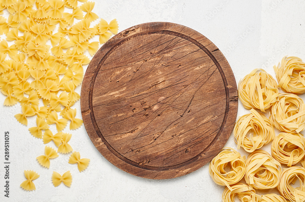 Round rustic wooden cutting board surrounded by different types of Italian pasta