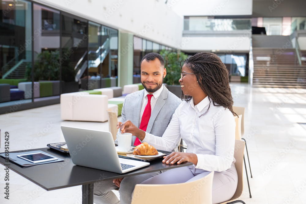 Obraz premium Happy business colleagues having breakfast in cafe. Business man and woman sitting at laptop outside and talking. Business breakfast concept