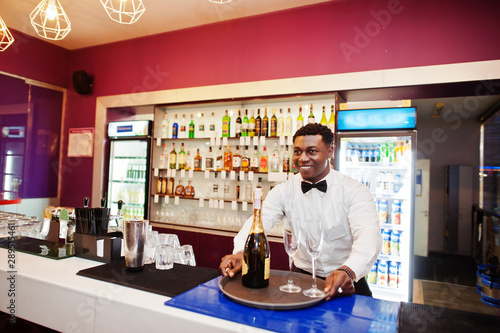 African american bartender at bar holding champagne with glasses on tray.