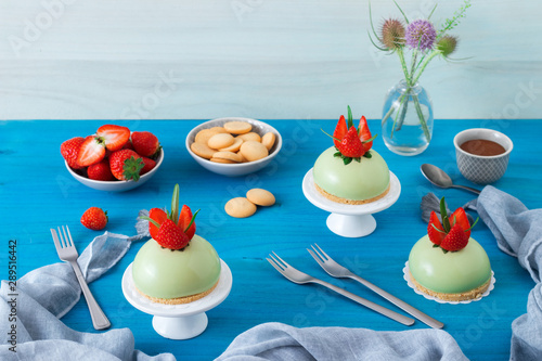 Blue wooden table with 3 no-bake strawberry mini sphere matcha cheesecakes with bowl of salt caramel and buiscuits, glass pitches and glass of milk.