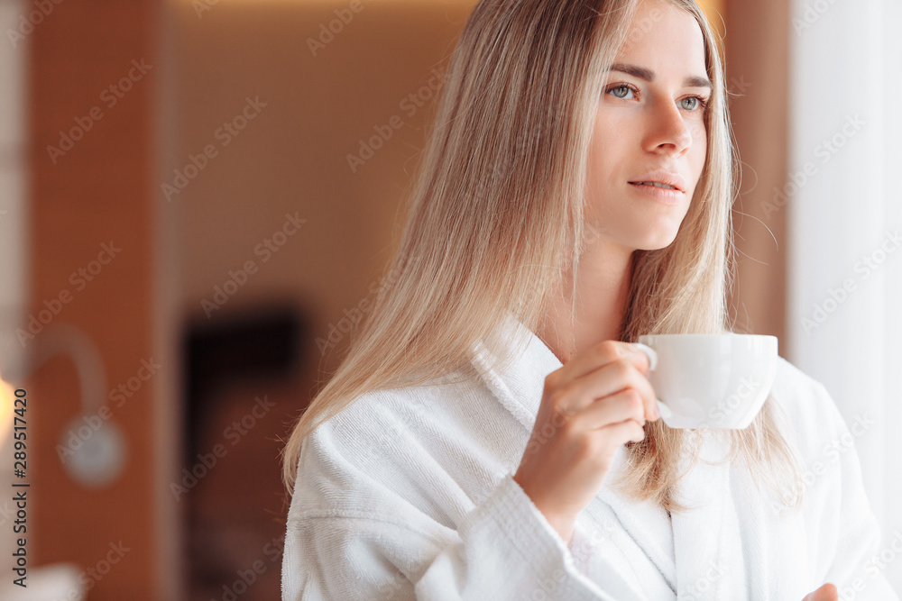 Beautiful young business girl is drinking coffee in the morning. Charming woman in a good mood starts the day with a smile, looks out the bedroom window. An invigorating drink helps set up for work Beautiful young business girl is drinking coffee in the morning. Charming woman in a good mood starts the day with a smile, looks out the bedroom window. An invigorating drink helps set up for work