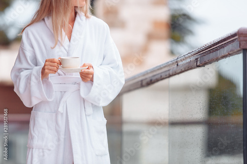 Charming healthy young blonde woman with a cup of coffee in a bathrobe walks barefoot on her large terrace enjoying the summer nature. Concept o vacation in own big country house or hotel