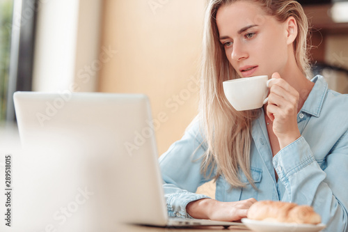 Slim focused young beautiful blonde woman checking social media messages while sitting at the kitchen table with a cup of coffee. Pretty woman checks orders in her online store. Copyspace