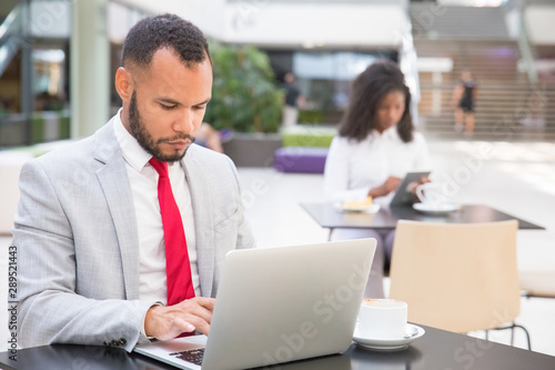 Focused serious businessman using laptop in co-working space. Young African American woman using digital device in background. Workspace concept