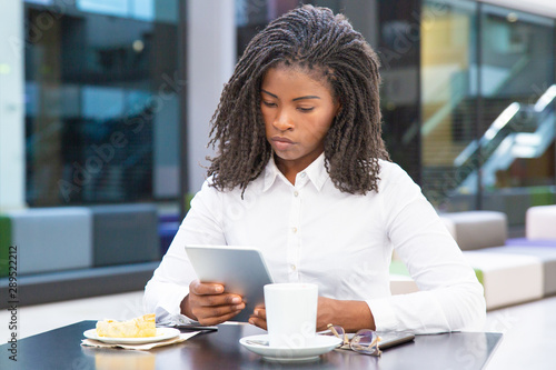 Serious student girl using tablet in cafe. Young African American woman sitting at table, holding gadget, looking at screen. Digital device concept