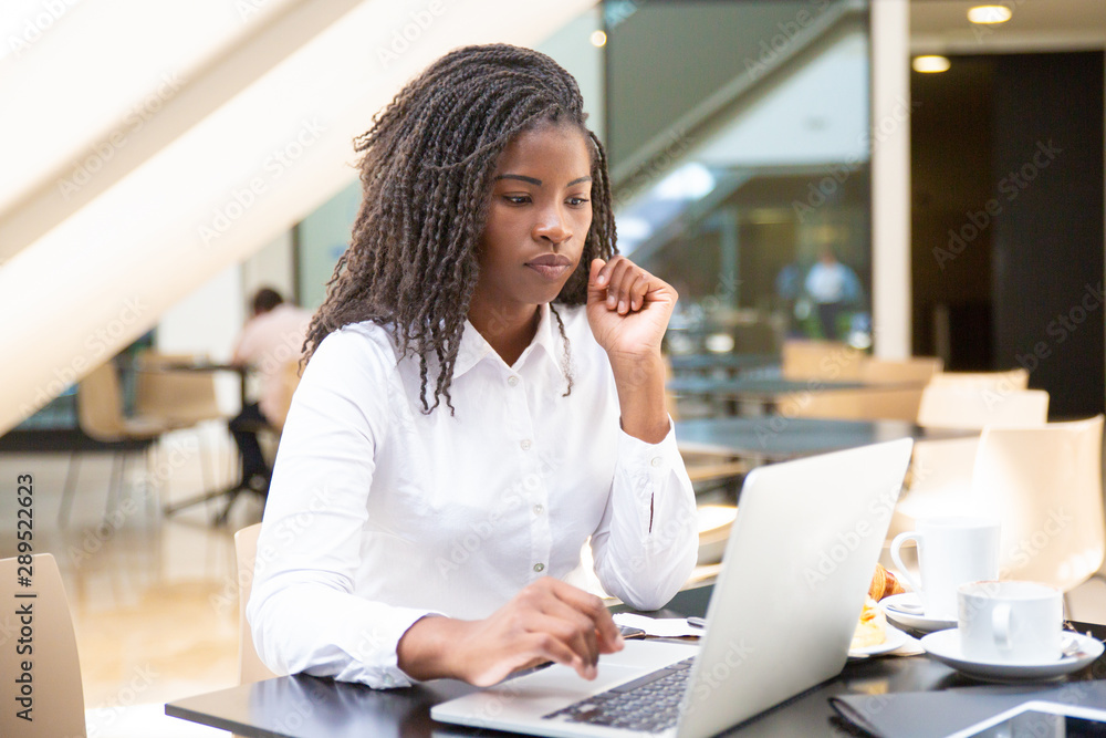 Focused female office worker using computer in coffee shop. African American business woman working on laptop in cafe and looking away. Internet technology concept Focused female office worker using computer in coffee shop. African American business woman working on laptop in cafe and looking away. Internet technology concept
