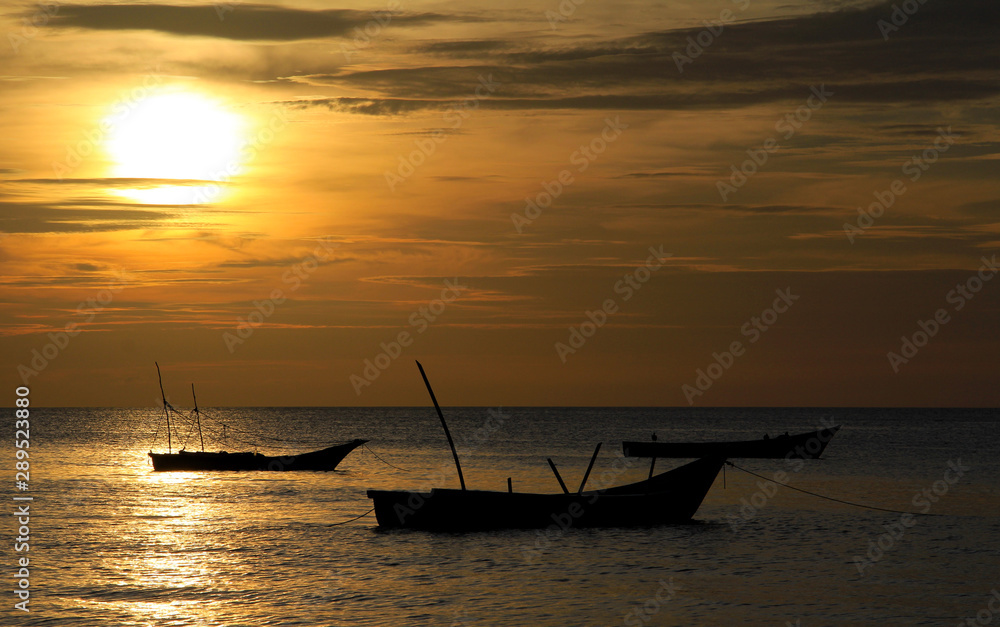 The sun that falls on the horizon backlights three boats in the sea.