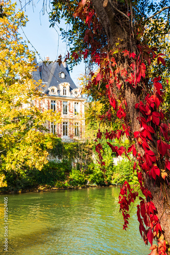 Beautiful autumn river landscape with red ivy and old house in L'Isle-Adam, Île-de-France, France. Colorful autumn nature background. Countryside tourism in autumnal season. Europe travel in fall.