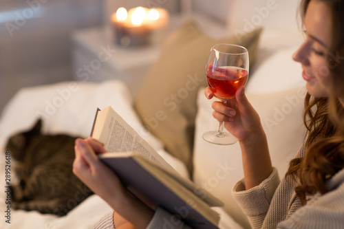 christmas, leisure and comfort concept - happy young woman reading book and drinking rose wine in bed at home bedroom