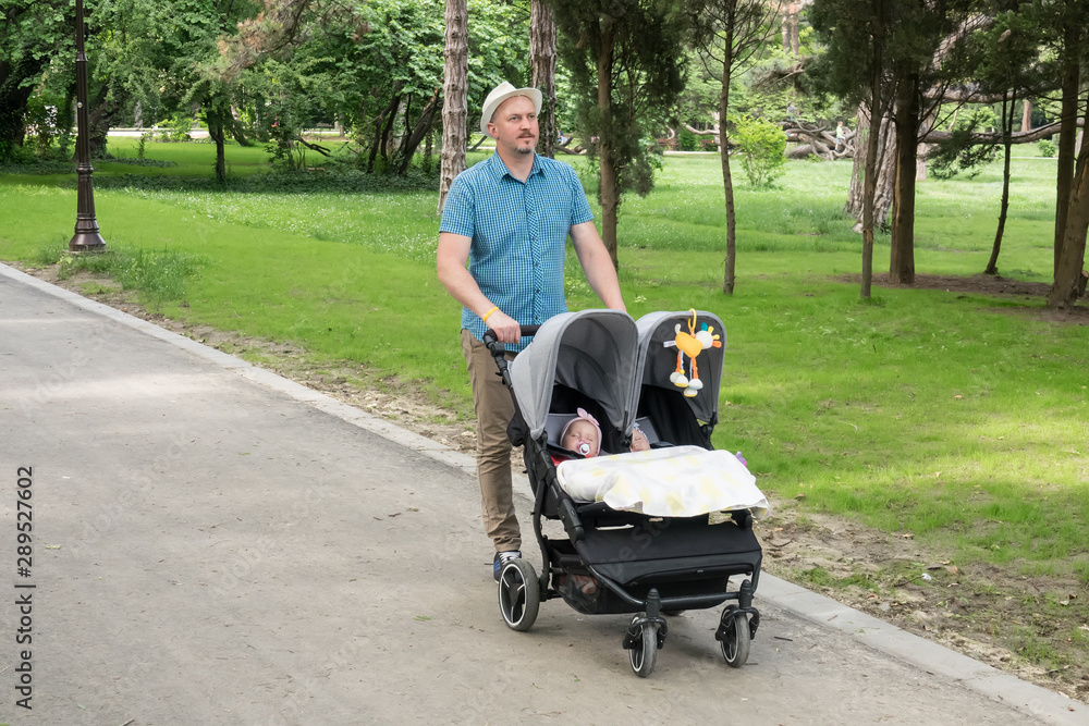 Father with kids in double stroller in a park. Man pushing twins stroller, pram. Father with kids in double stroller in a park. Man pushing twins stroller, pram.