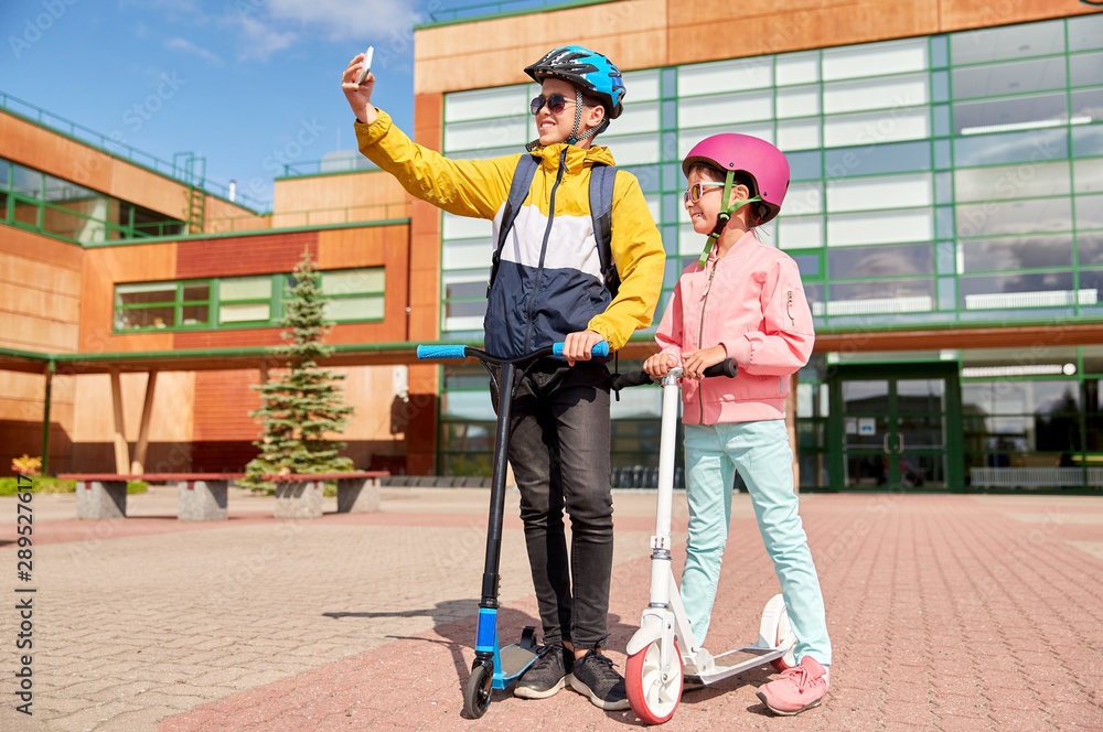 education, childhood and people concept - happy school children in helmets with backpacks and scooters taking selfie by smartphone outdoors