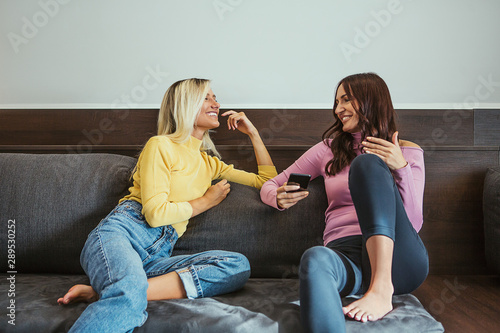 Two friends talking and laughing holding a phone sitting in a couch in the living room