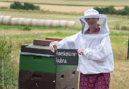 Imkerin vor zwei Bienenstöcken hält eine Kreidetafel mit dem Wort 