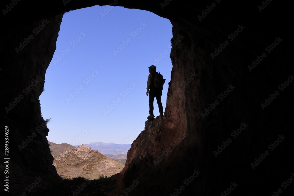 EXPLORER WITH HAT AND BACKPACK UP ON A SHELF IN A LARGE CAVE WATCHING A CASTLE IN THE DISTANCE