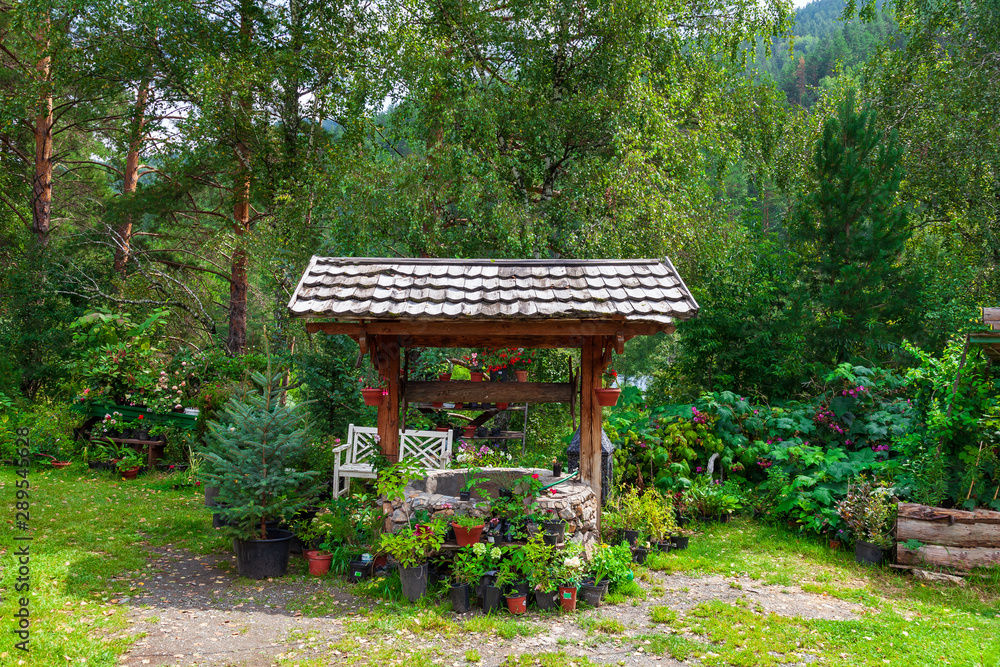 Naklejka premium Old retro stone well with a wooden roof with lots of flower pots and small trees in a botanical garden on a summer day. Green fresh plants on the nature.