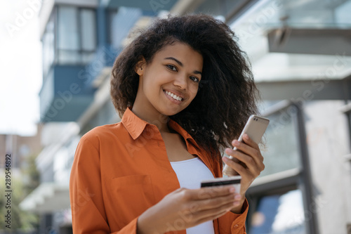 African American woman holding credit card, using smartphone with mobile application for online shopping with low prices and cash back. Happy hipster girl ordering food online, making transaction  photo