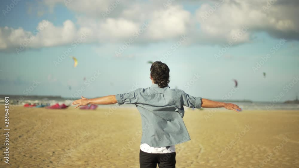 Young sensitive guy looking at kitesurfes flying in the sky under a blue and cloudy sky and opening arms by the sea. Beach in autumn or winter. Young sensitive guy looking at kitesurfes flying in the sky under a blue and cloudy sky and opening arms by the sea. Beach in autumn or winter.